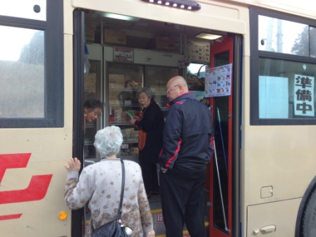 「準備中」と書かれた札の掛かかり、中には商品棚が見える移動販売のバスの出入り口に数人の高齢者が並んでいる様子の写真