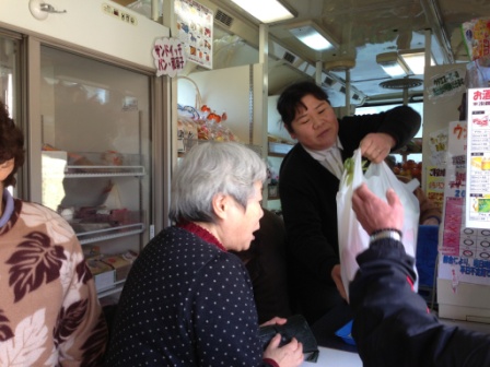 バスの中で買い物を終えた客に、女性店員が袋詰めした商品を手渡していている様子の写真