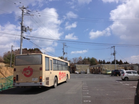 奥に仮設住宅の建物が見える駐車場にベージュ色の移動販売バスが停まっている写真
