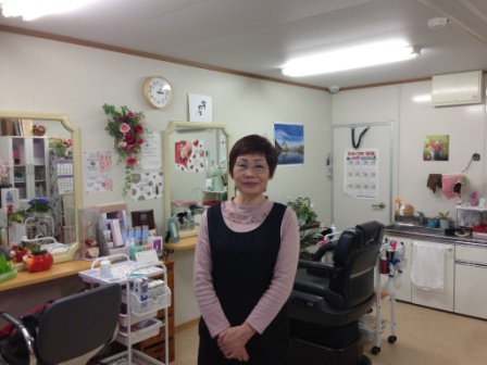 壁際に鏡が乗った台と椅子が並び、奥に流しがある美容室の店内に立っている山内さんの写真