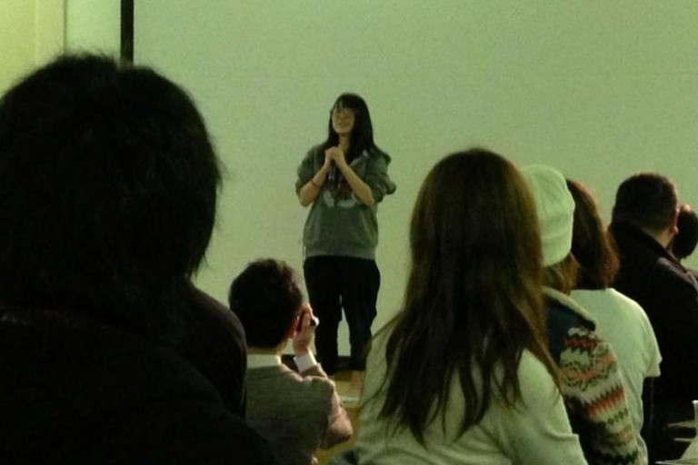 舞台の上で女子学生が両手でマイクを持ち語りかけるように話しており、暗い室内には彼女の話を静かに聞いている観客たちが椅子に座って並んでいる様子の写真
