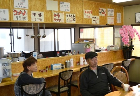 壁一面に手書きのメニューが貼られたカウンター席で、女性と店主の澤田力さんが席に座っている店内の様子を写した写真