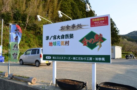 自然に囲まれた駐車場の横に「お食事処 津ノ宮大自然塾 地球元気村 金の鮫」と大きく書かれた看板が設置されている写真