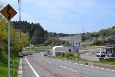 S字のカーブを示す標識が手前にある道路の右側に、民家や白いテントが点在し、左側には木々が多くある写真