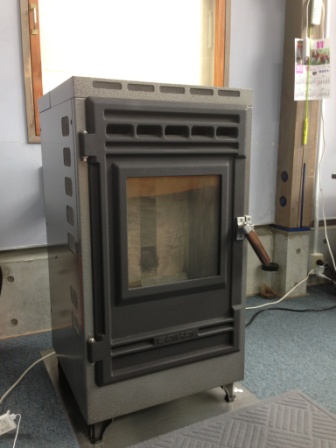 前面に大きな耐熱ガラスの窓と木製の取っ手がついたドアがある灰色の金属製ペレットストーブが室内に置かれている写真