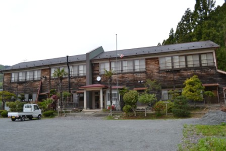 外壁が板張り木造の二階建ての古い校舎の前に植木がたくさん植えられている校舎の宿「さんさん館」の外観写真