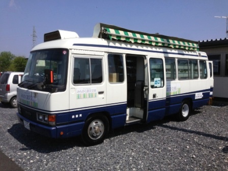 白と青のカラーリングの中型バスが駐車場に停車しており、車体上部には緑と白のストライプ柄の巻き取り式日除けが設置されていて、スライドドアが開いている状態の移動図書館の写真