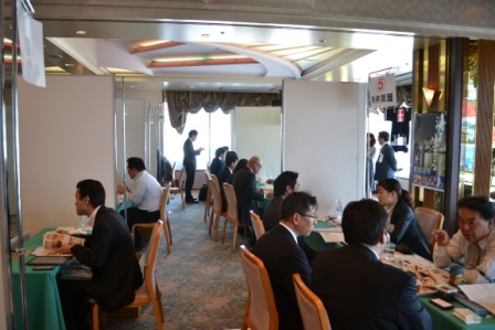 パーティションで区切られた会場内でスーツ姿の男女が数組に分かれて向かい合って座り、商品サンプルや資料を使いながら熱心に商談をしている様子を写した商談会場全体の写真