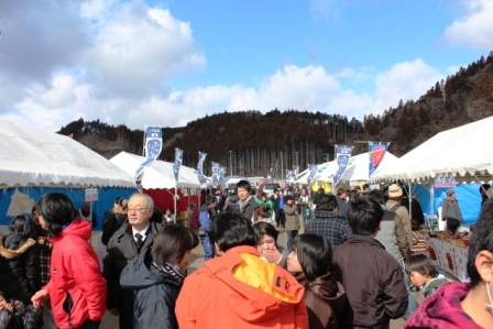 青空の下、白いテントの出店が両側に並び、たくさんの人々が冬の屋外でにぎやかに行き交いながら買い物や交流をしている福興市のイベントの様子を写した写真