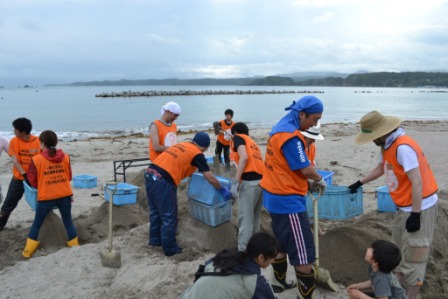 たくさんの人が3人一組で協力して青いかごを使ってふるいにかけながらがれきを取り除く作業をしている様子を写した写真