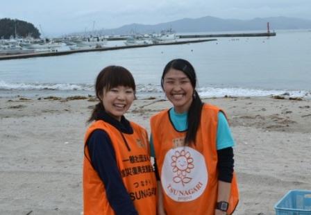 海辺の砂浜で、オレンジ色のビブスを着た2人の若い女性ボランティアが笑顔で並んで立ち写っている写真