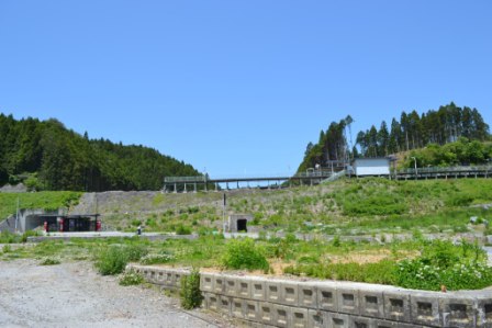 雲一つない晴れ渡った青空が広がり、奥には緑豊かな木々が生い茂った2つの丘が見え、斜面を上がった高台に白壁の小さな駅舎とホームが残っている旧歌津駅があり、左下に赤色の看板が設置された工事中のバス停が見える全体を撮影した写真
