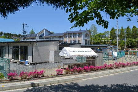 伊里前小学校の校庭に仮設住宅が並びテントが1つ張られ回りにピンク色の花が咲いている写真