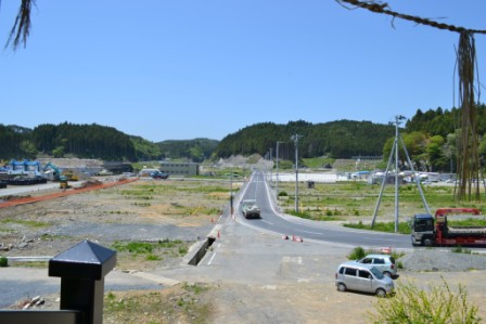 青空が広がる中、緑に覆われた山々に囲まれた広い平地に、一本の舗装された道路が真っ直ぐに伸びており、左側には重機が並べて停められている写真