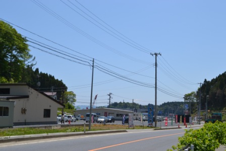 青空の下、道路際に建物やのぼり旗が立ち並び駐車場に車が複数停められている写真