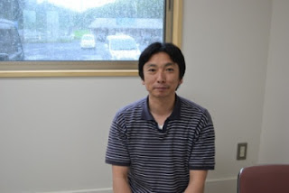 加工場内の窓際に座ってカメラを見つめるボーダーシャツの千葉孝浩さんの写真