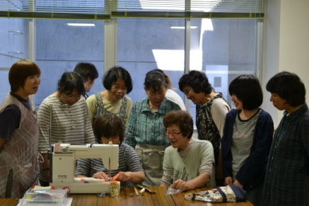 女性たちが机のまわりに集まり、1人の女性がミシンで縫い物をしている様子を他の人たちが興味深そうに見守っている写真