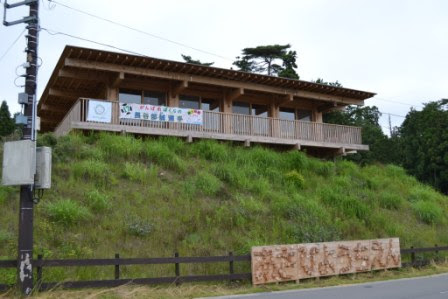 草の茂った小高い丘の上に木造の平屋建ての園舎が建っており、手すりのあるバルコニーの下に「あさひ幼稚園」の横断幕と木の看板が見える写真