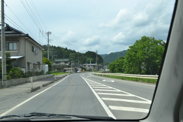 曇り空が広がる中、右にカーブした道路とその周囲の住宅、電柱、山々、木々が広がる風景を、車のフロントガラス越しに見た写真