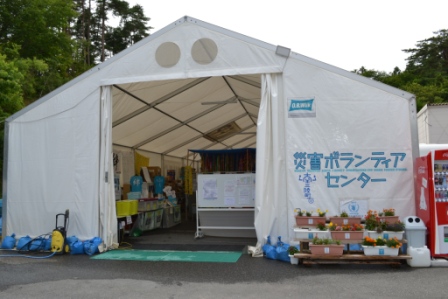白い大型テントの入口が開かれており、「災害ボランティアセンター」と青字で書かれた垂れ幕がかかり、入口には花のプランターや自動販売機が並んでいる写真