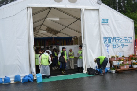 白い大型テントの「災害ボランティアセンター」の入口に複数のボランティアが集まっており、スタッフ用のベストを着て受付や準備をしている様子の写真