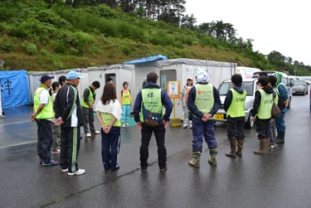 蛍光グリーンや黄色のスタッフベストを着たボランティアや作業員たちが、プレハブ近くに輪になって立ち集まり、作業開始前の打ち合わせをしている様子の写真