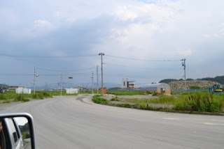 薄い雲が広がる青空の下、ビニールハウスや山積みにされた工事中の石の山が点在している様子を、運転席側から撮影した写真