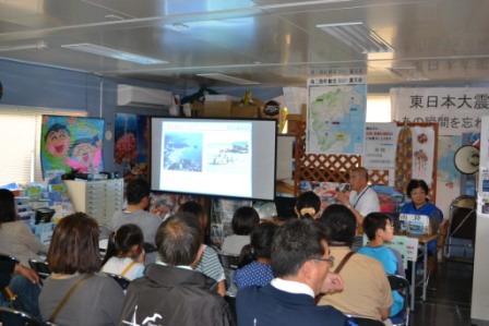 語り部の男性がマイクを持って話し、正面のスクリーンに写真を映し出しながら、多くの聴衆が真剣に耳を傾けている講話の様子が写っている写真