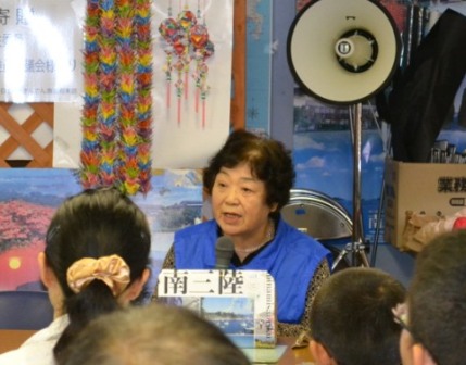 青いベストを着た高齢の女性菅原幸子さんがマイクを持ち、正面の子どもたちに向けて震災当時の体験を語っている講話の様子が写っている写真