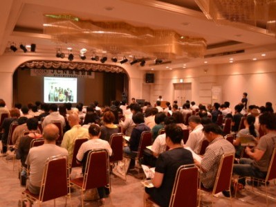 広い会場で多くの人が椅子に座り、前方のスクリーンに映し出されたスライドを見ながら、現地会議に参加している様子の写真