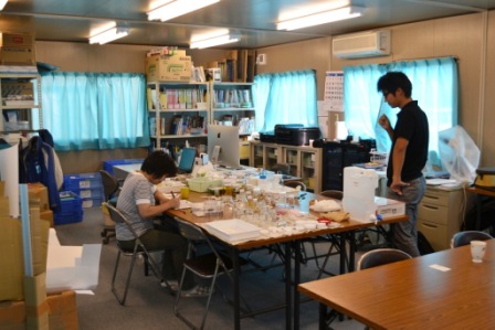 プレハブの室内の中央に長机が向かい合うように並べられ、机の上には標本整理に使う瓶や資料が広げられ、女性が座って作業し、黒田和真さんが立って様子を見ており、周囲には資料が並んだ本棚があるネイチャーセンター準備室の室内の様子の写真