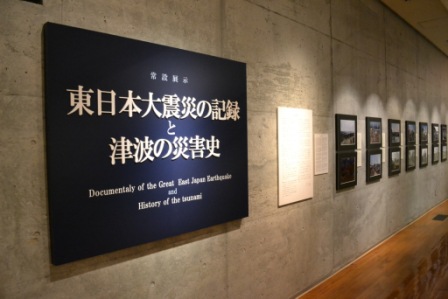 白い文字で『東日本大震災の記録と津波の災害史』と日本語で書かれている黒い案内板が、コンクリートの壁に取り付けられており、その奥に写真が並んでいる展示室入り口の案内板を写した写真