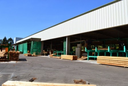 大型の屋根付き製材工場の外観で、建物の中には緑色の製材機が並び、屋外にはカットされた木材が整然と積まれている様子が見える製材工場の写真