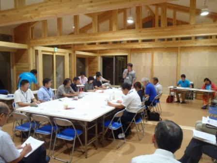 会議室で、参加者の男女が長机を囲んで座り、真剣な表情で資料を見ながら地域づくりに関する話し合いを行っている様子の写真