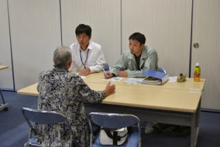 会場内の長机を挟んで、書類を前にした白いシャツの男性職員と緑のジャンパーの男性職員が、背を向けた高齢の女性住民と対面して相談に応じている地域相談会の現場の写真