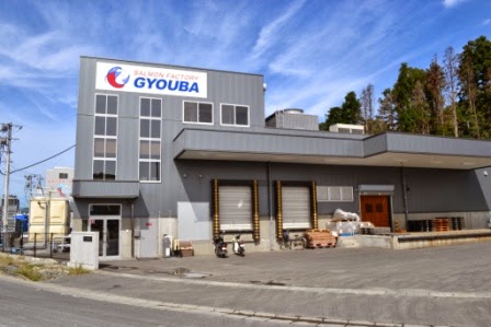 「SALMON FACTORY GYOUBA」と書かれた看板が掲げられたグレーの水産加工場の建物正面に、トラックの搬入口や資材が見える外観写真
