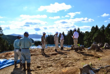 伐採された山の上で青いビニールシートが地面に敷かれた現場に、ヘルメットを着用した関係者たちが集まり、発掘調査視察が行われている場所を視察している写真