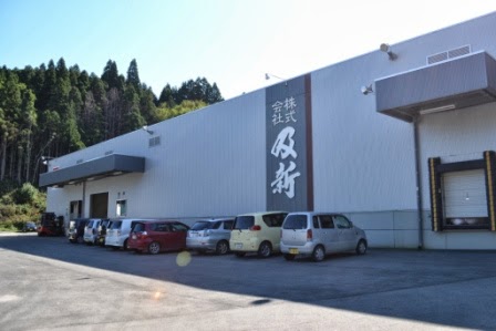 工場の外壁に「株式会社 及新」と書かれた大きな看板があり、建物の前には自動車が数台停まっている、山あいに建つ水産加工会社の写真
