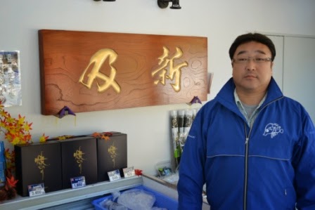 木目のある茶色い板に金色で「及新」と立体的に彫られた社名看板の横に立つ及川社長の写真