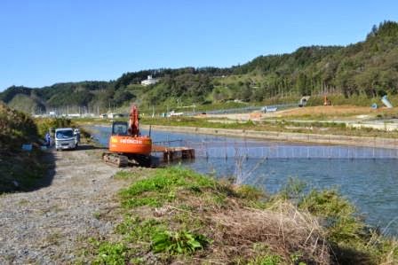 川沿いに重機と作業車が停まり、川の中にはネットの仕切りが設置されている鮭の捕獲施設の設置工事現場の写真