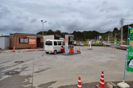 曇り空の下、広い敷地の中に1台だけ給油機がある仮設店舗で白い軽自動車が給油している様子の写真