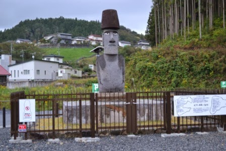 震災復興支援のシンボルとしてチリから贈られた本物のモアイ像が、フェンスに囲まれた広場に立ち、背景には南三陸町の住宅地と山の斜面が見える写真