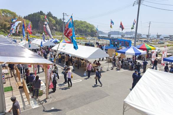 カラフルな旗やのぼりが風になびき、白いテントが並び、多くの方々で賑わっている伊里前福幸商店街の写真