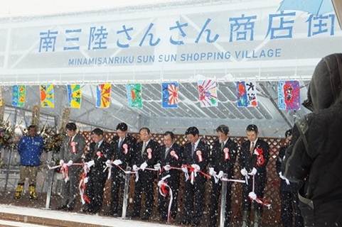 雪が降る中、スーツ姿の関係者たちが「南三陸さんさん商店街」と書かれた大きなテントの下に並んでテープカットを行っている様子の写真