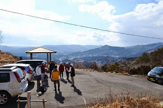 山並みを見渡す景色の駐車場に数台の車が停まっており、帽子をかぶった人々が輪になって集まっている様子の写真
