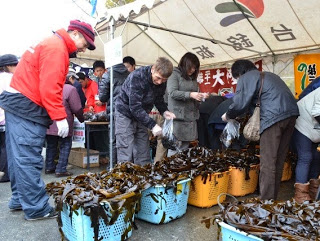 オレンジや青のコンテナにたくさんの生わかめが入れられ、たくさんの人たちが買いに来ている様子を写した写真