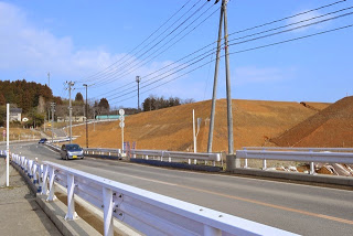 青空の下、車が走る道路の右側に盛土された茶色い土の斜面が広がっている様子の写真
