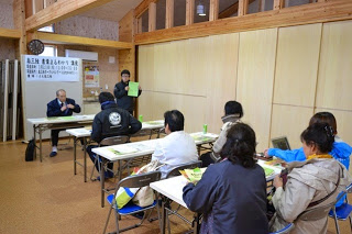 長机が3つ1列に並べられ、参加者がその机に座って前方の関係者の説明を真剣に聞いている様子の写真