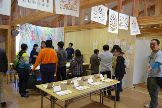 部屋には木製の柱や梁があり、天井からは切り絵のきりこの作品が吊るされ、複数の人々が展示物を見たり、中央にはテーブルがあり、その上にはいくつかの作品が展示されている室内の写真