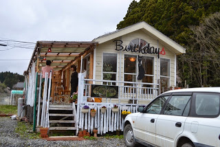 木製のデッキと階段がついた白い建物で、窓の上に「Birthdays」と書かれた看板が掲げられており、入り口付近には二人の人物が立っているカフェの外観写真